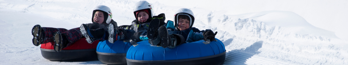 Three individuals wearing helmets and winter clothing, sitting in inflatable snow tubes on a snowy surface. The tubes are blue and red.