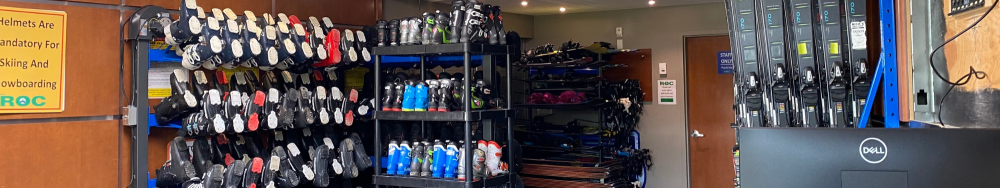 Alt text: A room filled with various types of sports equipment, primarily focusing on skiing and snowboarding gear. Shelves on the left side are stacked with ski boots, while the center has racks holding multiple pairs of skis and snowboards. The right side has more skis leaning against the wall. A sign on the left reads: "Wellness Av mandatory for Skiing And Snowboarding." A Dell computer monitor is visible in the bottom right corner.