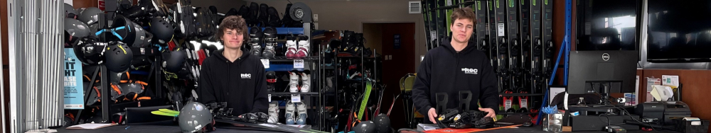 Two individuals in a store or workshop setting, likely related to sports equipment. The background is filled with various items such as helmets, shoes, and other gear. The individuals are standing behind a counter with more equipment on it.